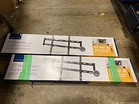 Two boxed Insignia TV wall mounts stacked on a floor showing front of boxes with brand and description.