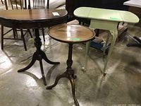 Photo showing all three tables together: a dark wood demilune table, round wood pedestal table, and painted green drop-leaf table with carved legs.