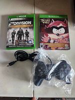 Two Xbox One game cases: Tom Clancy's The Division Gold Edition and South Park, next to a black wired Xbox One controller in plastic bag and a USB cable.