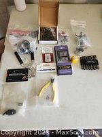 Photo showing assorted hand tools arranged on a table including various bits, pliers with yellow handles, boxed parts, and small plastic bags with hardware.