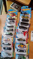 Photo showing the majority of Hot Wheels cars sealed in their original plastic and card packaging. Cars vary in design, color, and model style, laid out for visual confirmation of quantity and condition.