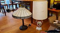Both lamps positioned on floor showing full body and shade of ceramic floral urn lamp with fabric shade and stained glass style lamp with geometric cream and brown panels.