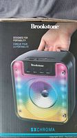 Front and top panel of Brookstone SXChroma Bluetooth speaker showing multicolor LED light grill, control buttons, and branding.