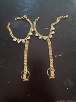 Two gold tone hand pieces laid flat on a dark surface showing chains with clear crystals and attached rings