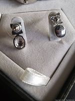 Close up of two pairs of silver-tone stud earrings, one oval textured and one floral and round dome design, displayed on clear stands on soft fabric.