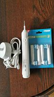 White Oral-B electric toothbrush handle next to charger adapter with power cord and pack of four replacement brush heads in original packaging on wooden surface.