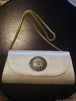 Front view of small cream purse with gold-tone round studded clasp and gold-tone chain strap laid on black surface.