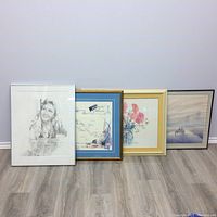Photo showing four framed artworks leaning against wall on floor: pencil portrait, nautical map, floral painting, lakeside scene
