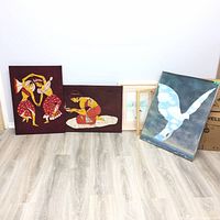 Collection of five artworks leaning against a wall, including two distinctive burgundy cloth paintings and a framed bird silhouette art piece with a cloudy sky background, plus two smaller framed lighter artworks.