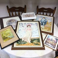 Variety of framed artwork pieces placed on and around two wooden chairs and a table, showing a central large painting of a woman in a pink dress and several surrounding paintings with various subjects including a man, landscapes, and floral piece.