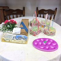 Overview photo showing Easter baskets with decorative filler, Avon hand steamer in box, purple egg holder, and artificial flowers on table.