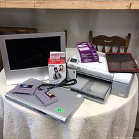 Overall electronics and gaming collection on table: Sony CRT monitor, Epson printer with cables, VTech cordless phone in box, Philips DVD player, red tablet case and two SNES cartridges on DVD player