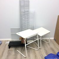 Two small white adjustable TV tables, black wooden computer shelf and white metal wire closet shelving panels in corner against wall on laminate flooring.