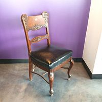 Side view of vintage wooden dining chair showing carved details on backrest, upholstered leather black seat, and curved front leg with wheel.