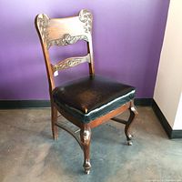 Front-left view of vintage wooden dining chair showing carved backrest, black leather padded seat, curved legs, and front caster wheels.