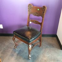 Front left angle view of the vintage carved mahogany chair with black leather seat and caster wheels on front legs.