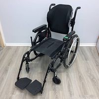Front left angled view of the wheelchair showing the cushioned backrest, missing seat pad, armrests, and footrests.