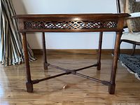 Full side view of wooden accent table showing carved lattice work on apron and cross stretcher base with turned legs.