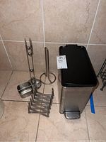 Photo showing all items included in the lot: metal trash can with black lid, accordion rack, paper towel holder, small container and vertical stand with three small bells.