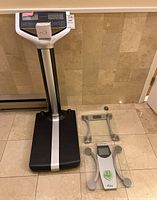 Three bathroom scales arranged on tiled floor: Accuro DBW100 digital scale with vertical display panel, and two glass digital scales from Weight Waters and Taylor brands.