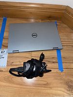 Closed Dell silver laptop with Dell logo on top and power cord charger coiled beside it on wooden floor.