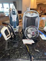 Image of two electric irons, a portable heater, and small desk fan arranged on a table.