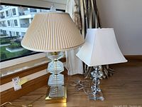 Two table lamps placed on wooden windowsill. One with pleated beige fabric shade and stacked geometric glass base on gold-tone platform. Second with white rectangular shade and clear glass base.