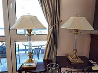 Two tall table lamps with ribbed glass and brass columns on square bases with white fabric shades and crystal pull chains, placed on furniture against a wall near a window.