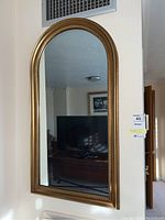 Wall-mounted rectangular mirror with arched top showing reflection of room and furniture, brass toned detailed frame visible.