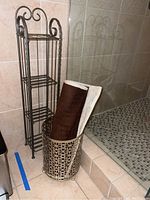 Metal waste basket with decorative cutout pattern holding folded brown and white bath mat beside shelving unit