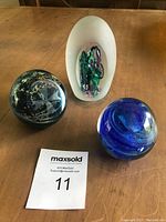 Three art glass paperweights on wooden surface including frosted glass with colorful internal design, dark greenish cross-etched piece, and blue swirling glass piece.