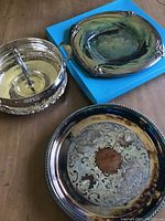 Three assorted silver and silver-plated serving trays/platters displayed on a table, one on a blue box