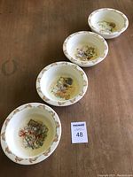 Four white bone china bowls with different Bunnykins story scenes inside and border of running bunnies around rim, arranged in a row on a wooden surface.