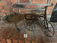 Cast iron bird bath and wrought iron tricycle plant holder positioned on brick floor in front of a brick wall.