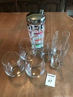 Vintage mid century cocktail shaker surrounded by six drinking glasses including three stemless wine glasses and three high ball glasses on wooden table.