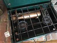 Top view of the camping stove showing black grilling bars, fuel tank, and burners inside green metal case.