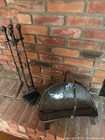Full view of the forged metal fireplace tool set with stand and tools next to the brick hearth, showing poker, tongs, shovel, broom with twisted metal handles.