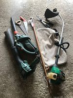 View of all garden tools lying on concrete floor showing leaf blower, vacuum bag, weed whacker, hand saws, branch trimmer, and hedge trimmers.