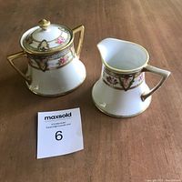 Top-down view of sugar bowl with lid and creamer jug, showing hand-painted roses and gold accents, placed on wooden surface next to auction lot number