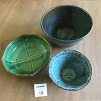 Three studio pottery bowls displayed on a wooden surface: one green octagonal, one marbled blue-grey, one large blue bowl.