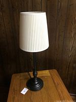 Tall black table lamp with white pleated fabric shade on wooden table against wood panel background.