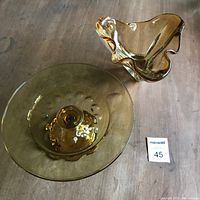 Two amber glass pieces displayed on a wooden surface: a round cake pedestal dish and a ruffled edged candy bowl, showing size and shape contrast.