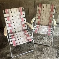 Two vintage 1960s folding lawn chairs with silver metal tubular frames, white and pink woven webbing seats and backs, and off-white plastic armrests.