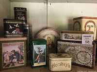 Front view of assortment of vintage metal biscuit and cookie tins showing different shapes, colors, and designs including Beatrix Potter, Hershey's, and floral patterns.