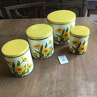 Four vintage tin canisters with yellow daisy floral design on white background and yellow lids displayed on wood table.