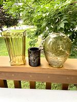 Three items shown on a wooden railing outdoors: two amber glass vases and one dark art glass pottery candle holder. The tall rectangular amber vase has vertical ribs and a flared opening. The oval amber vase has a cracked glass effect. The candle holder is dark and cylindrical with mottled texture.