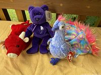 Three Beanie Babies together on wooden bench including the Princess Diana bear, red bull, and blue peacock with colorful tail feathers.
