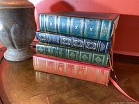 Four hardcover books with decorative spine designs stacked horizontally next to a vase against a red wall