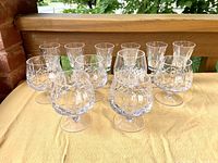 Front angle showing five smaller crystal glasses with faceted geometric cuts and short stems along with five larger snifter-style crystal glasses, all on a yellow surface outdoors.