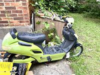 Side view of the green electric scooter with signs of wear and water droplets on seat, parked on concrete near brick wall and garden area.